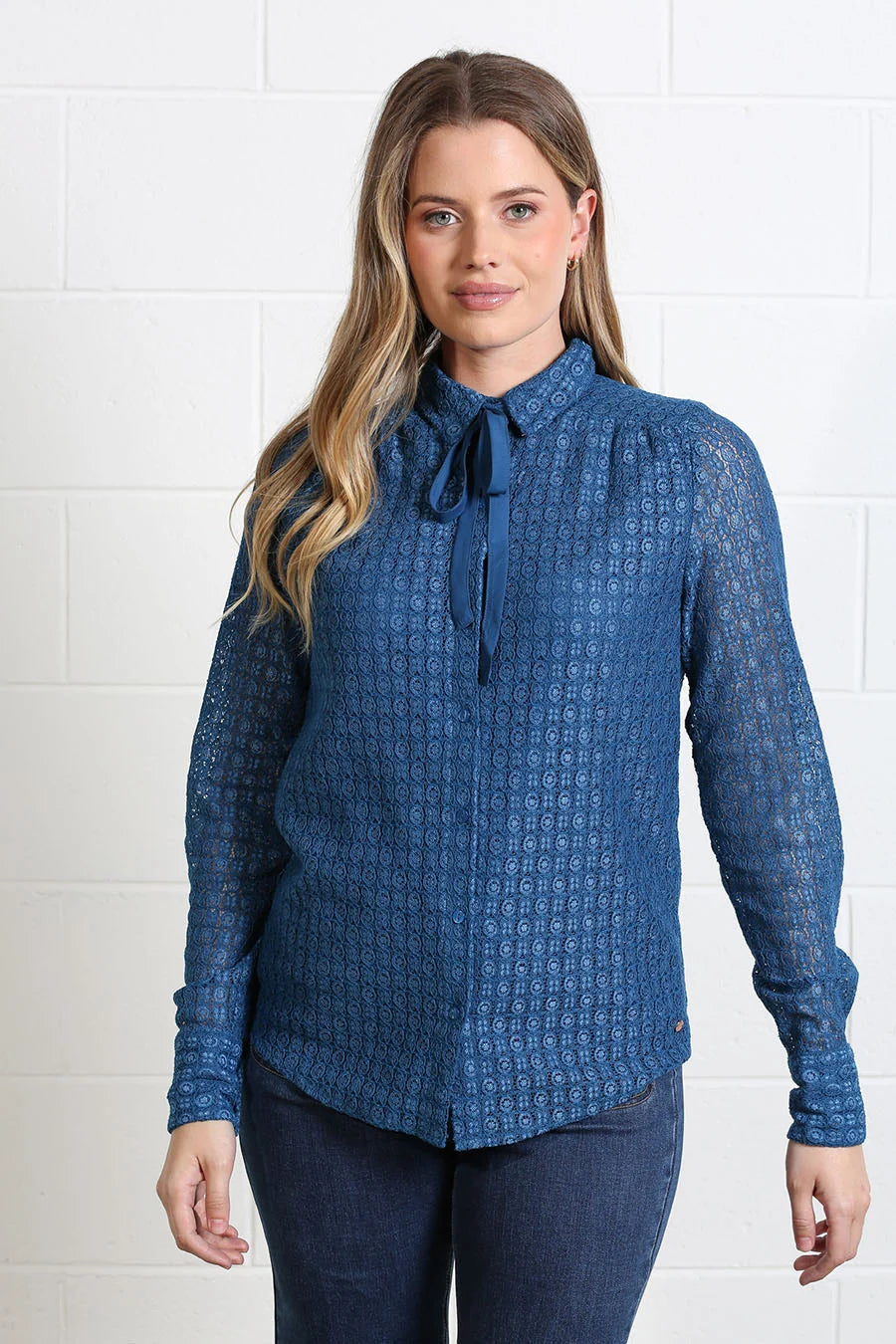 A woman modeling a blue patterned long-sleeve blouse with a tie-neck detail, paired with dark blue jeans, standing against a white brick wall.