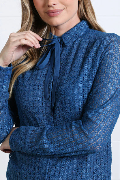 A woman wearing a blue lace long-sleeve blouse with a tie-neck detail, shown in a close-up shot against a white paneled wall.