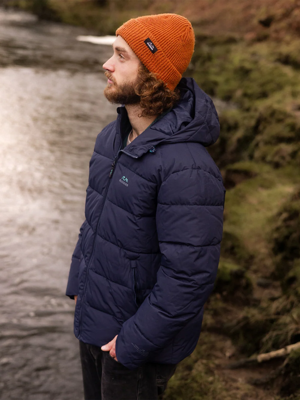 Man wearing a blue puffer jacket and orange beanie standing by a river