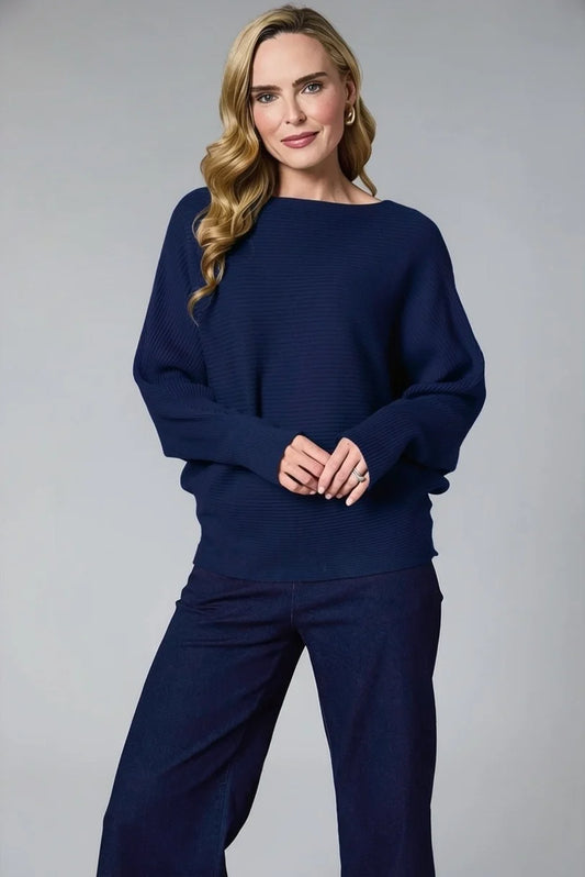 A woman modeling a navy blue ribbed knit sweater with a wide boat neckline and long sleeves, paired with matching navy wide-leg trousers, against a solid gray studio background.