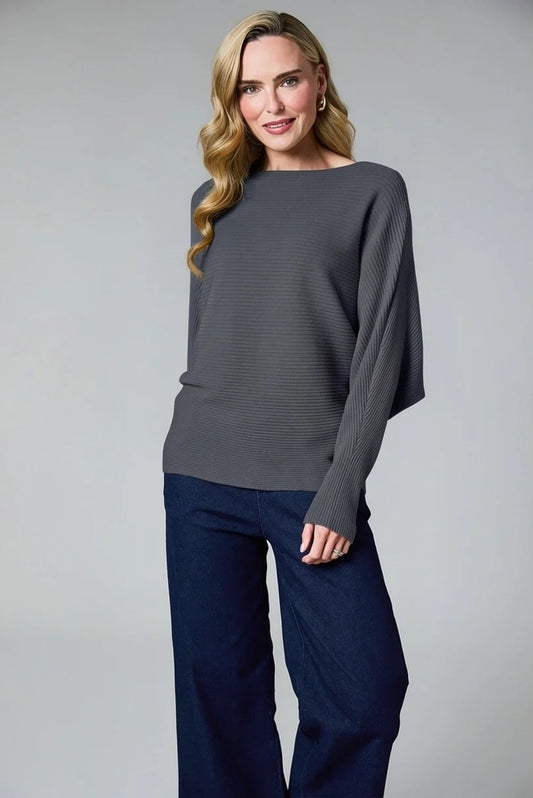 A woman modeling a dark gray ribbed knit sweater with draped sleeves and dark navy wide-leg pants, photographed against a neutral gray studio background.