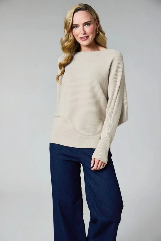 A woman modeling a cream ribbed knit sweater with wide sleeves and dark navy wide-leg pants, photographed against a neutral light gray studio background.