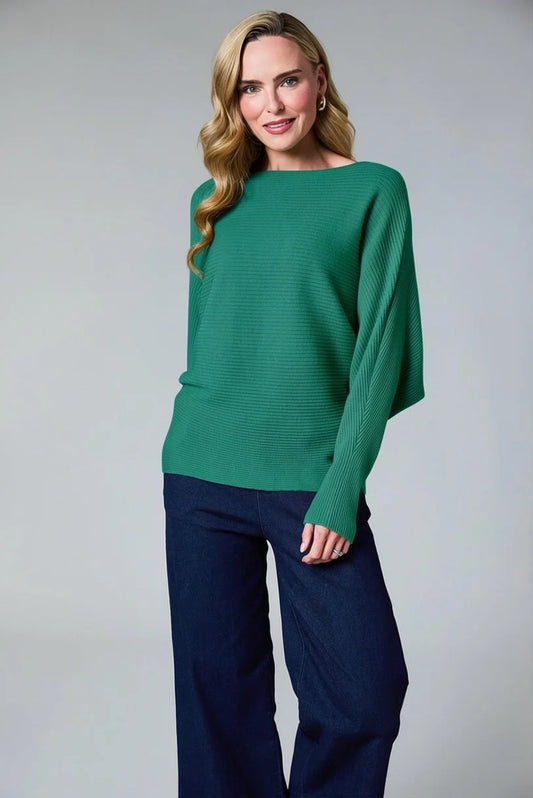 A woman modeling a green ribbed knit sweater with long draped sleeves, paired with dark navy wide-leg pants, against a neutral gray studio background.