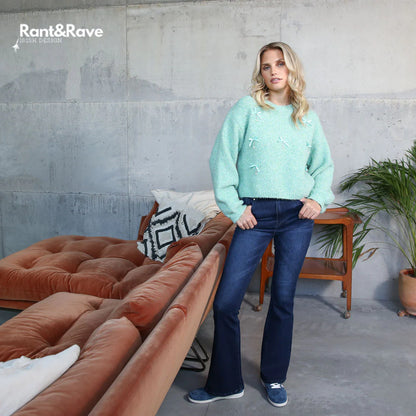 Woman in a light blue sweater and dark jeans standing in a modern living room with a brown sectional sofa.