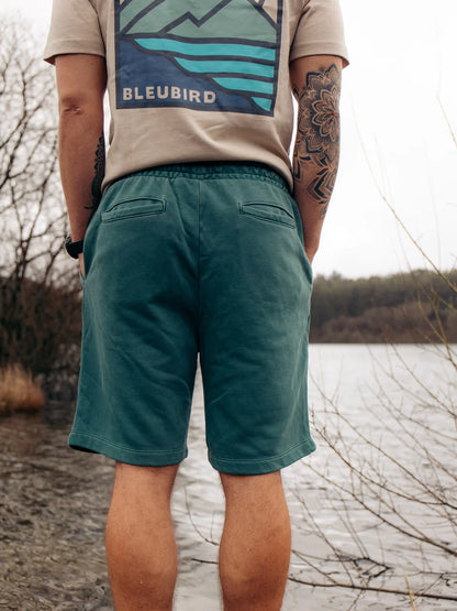 Person wearing teal shorts and a beige shirt with a bluebird logo by a body of water.