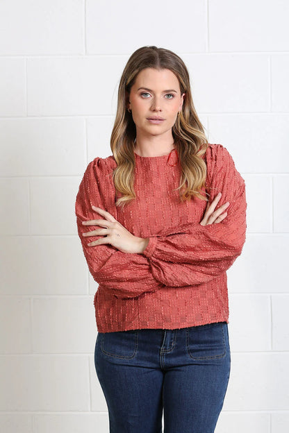 A woman with blonde hair stands with arms crossed, wearing a textured, rust-colored long-sleeve blouse and dark blue jeans, posing against a white brick wall.