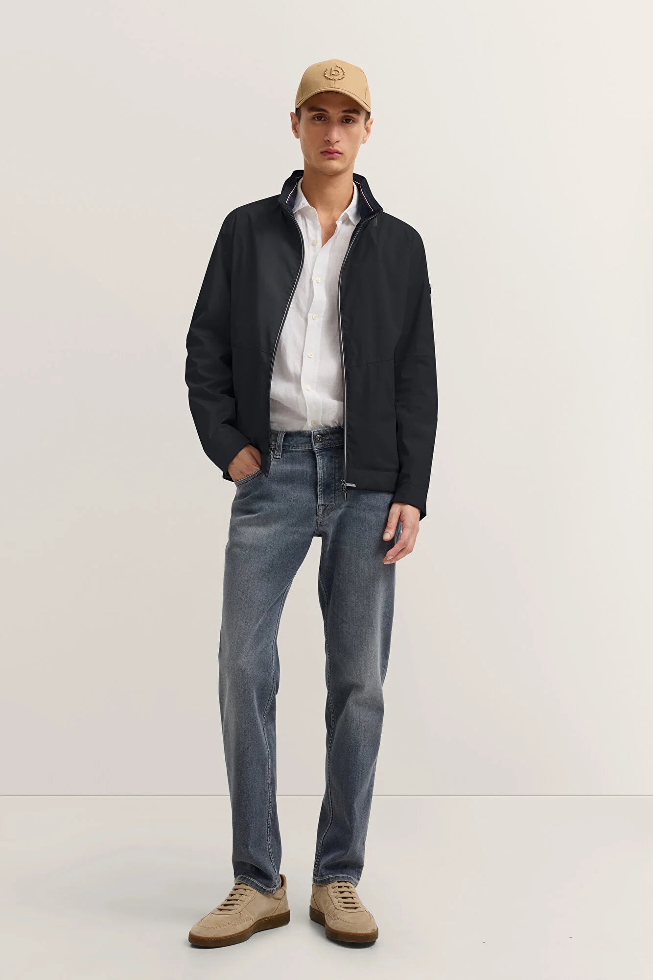 Man wearing a black jacket, white shirt, blue jeans, and beige cap on a white background