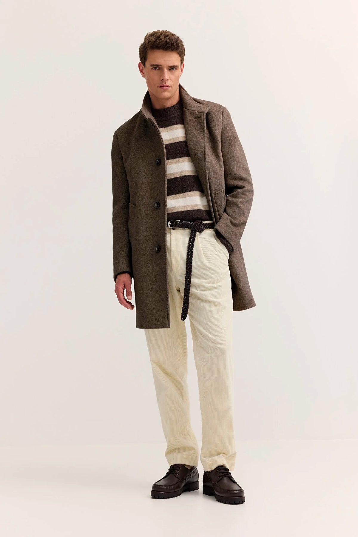 A male model stands against a plain light studio background wearing a brown overcoat over a black-and-beige striped sweater, cream trousers, and dark brown shoes.