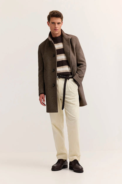 A male model stands against a plain light studio background wearing a brown overcoat over a black-and-beige striped sweater, cream trousers, and dark brown shoes.