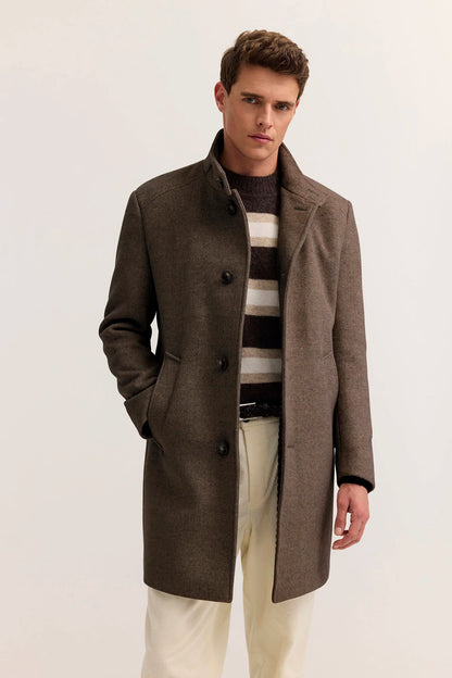A man wearing a mid-length brown wool-blend coat over a striped sweater and cream trousers, standing against a plain light background.
