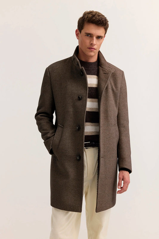 A man wearing a mid-length brown wool-blend coat over a striped sweater and cream trousers, standing against a plain light background.