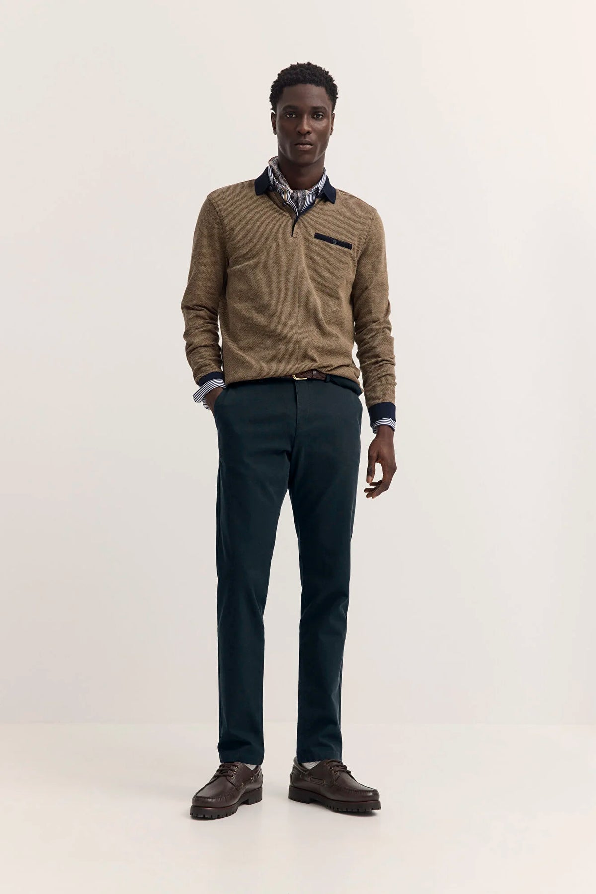 A man modeling a tan quarter-zip knit sweater over a collared shirt, paired with dark navy trousers and brown leather shoes, standing against a plain off-white studio background.