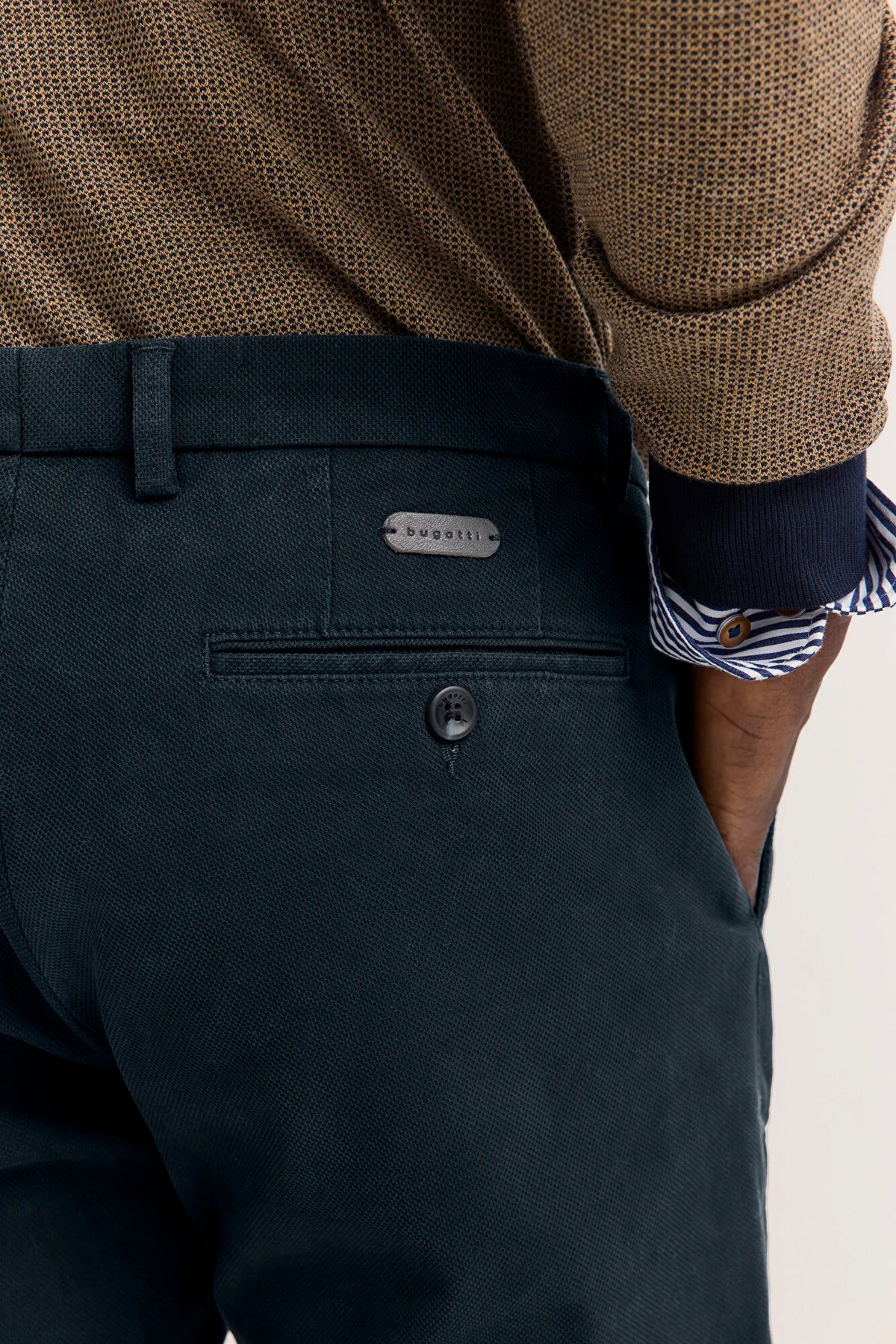 Close-up of a person wearing dark teal trousers, a brown textured sweater, and a striped shirt cuff, showing the back pocket and a small silver-tone brand plaque.