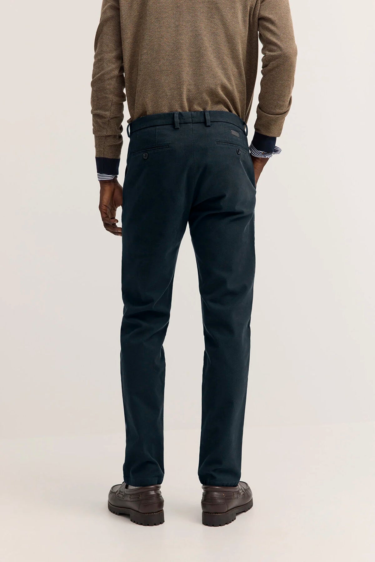 A man viewed from the back wearing dark navy trousers, a brown knit sweater, and brown leather shoes, standing against a plain light gray studio background.