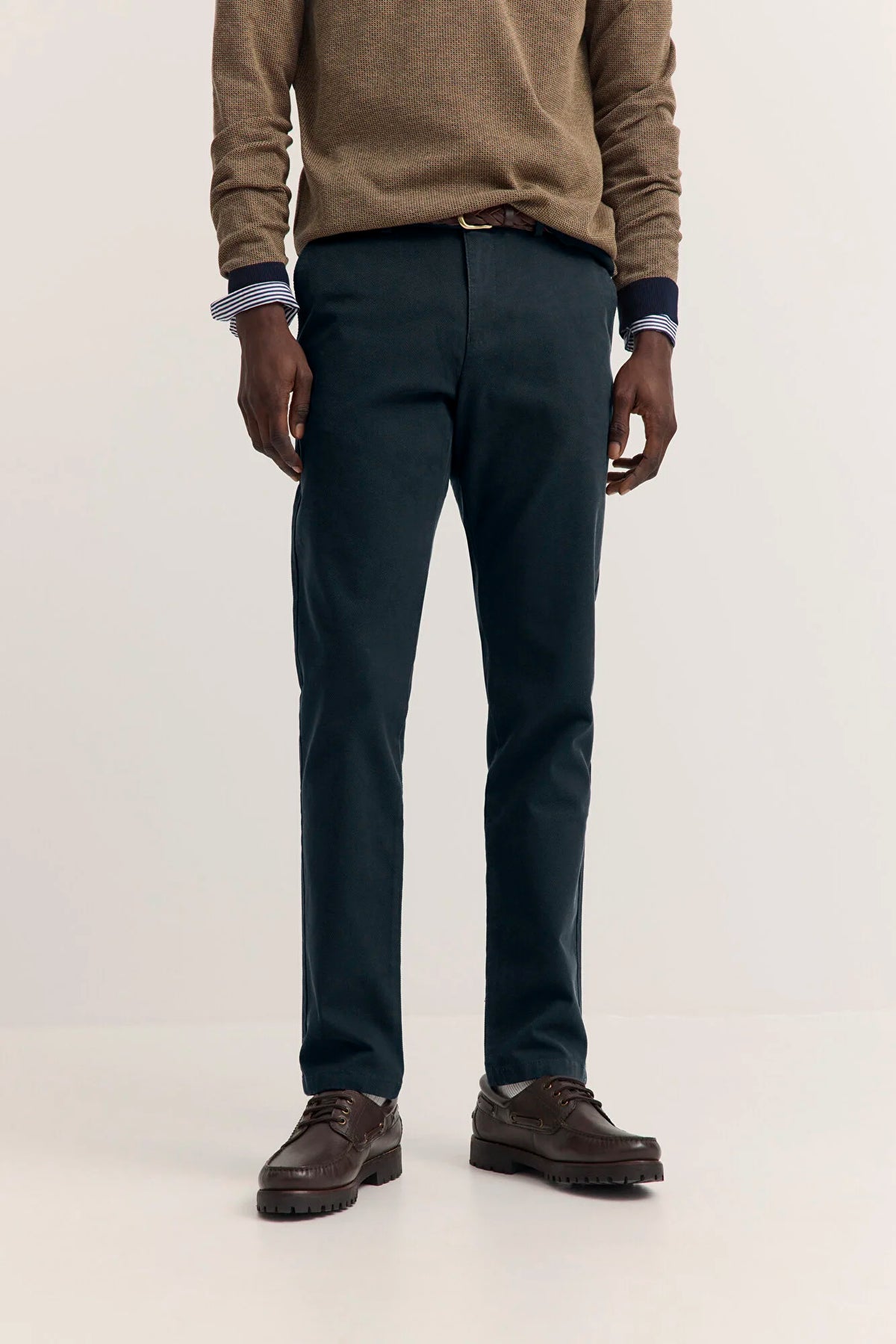 A man wearing dark teal slim-fit trousers, a brown knit sweater, and brown leather shoes, shown from the chest down against a plain light background.
