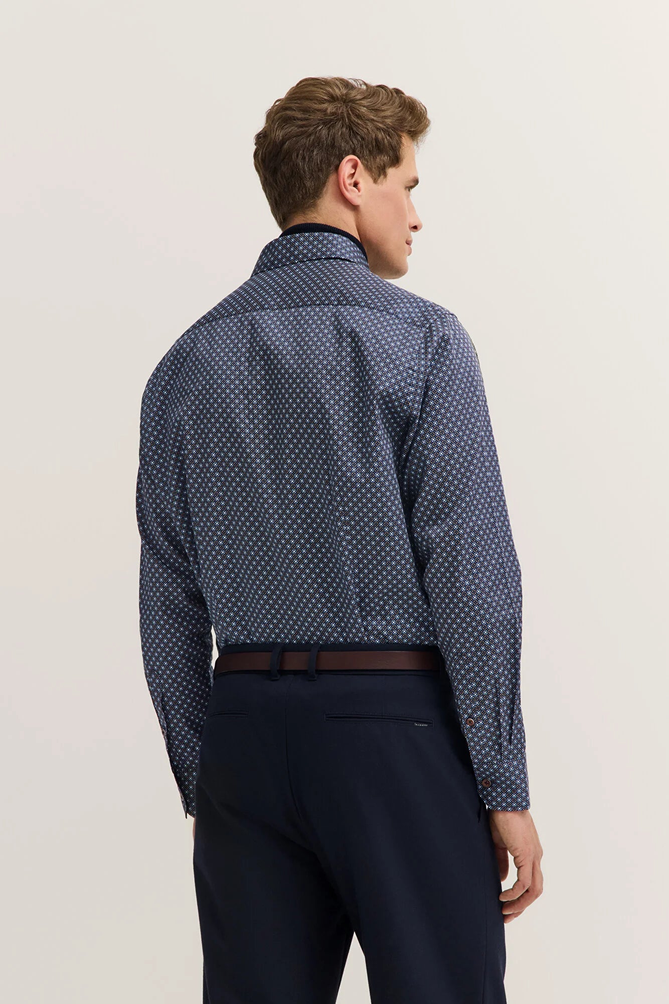 Man wearing a patterned blue shirt and dark pants on a light background