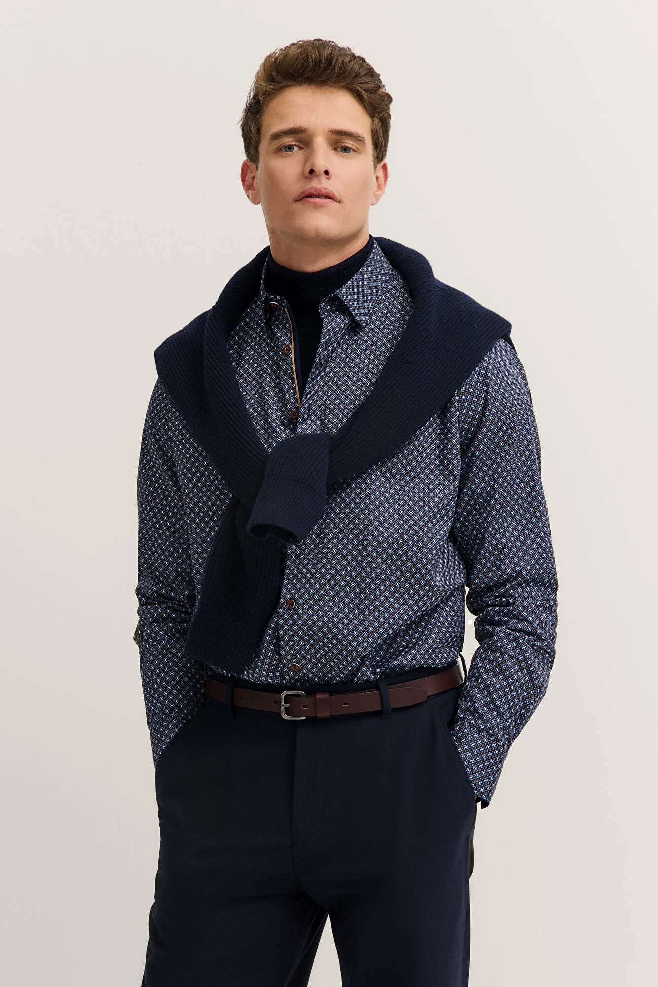 Man wearing a patterned shirt with a navy sweater over it on a plain background