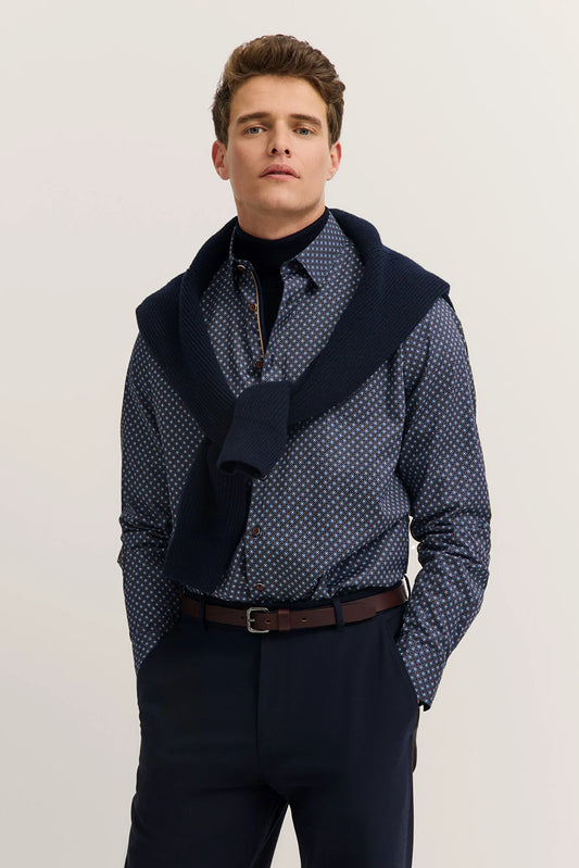 Man wearing a patterned shirt with a navy sweater over it on a plain background