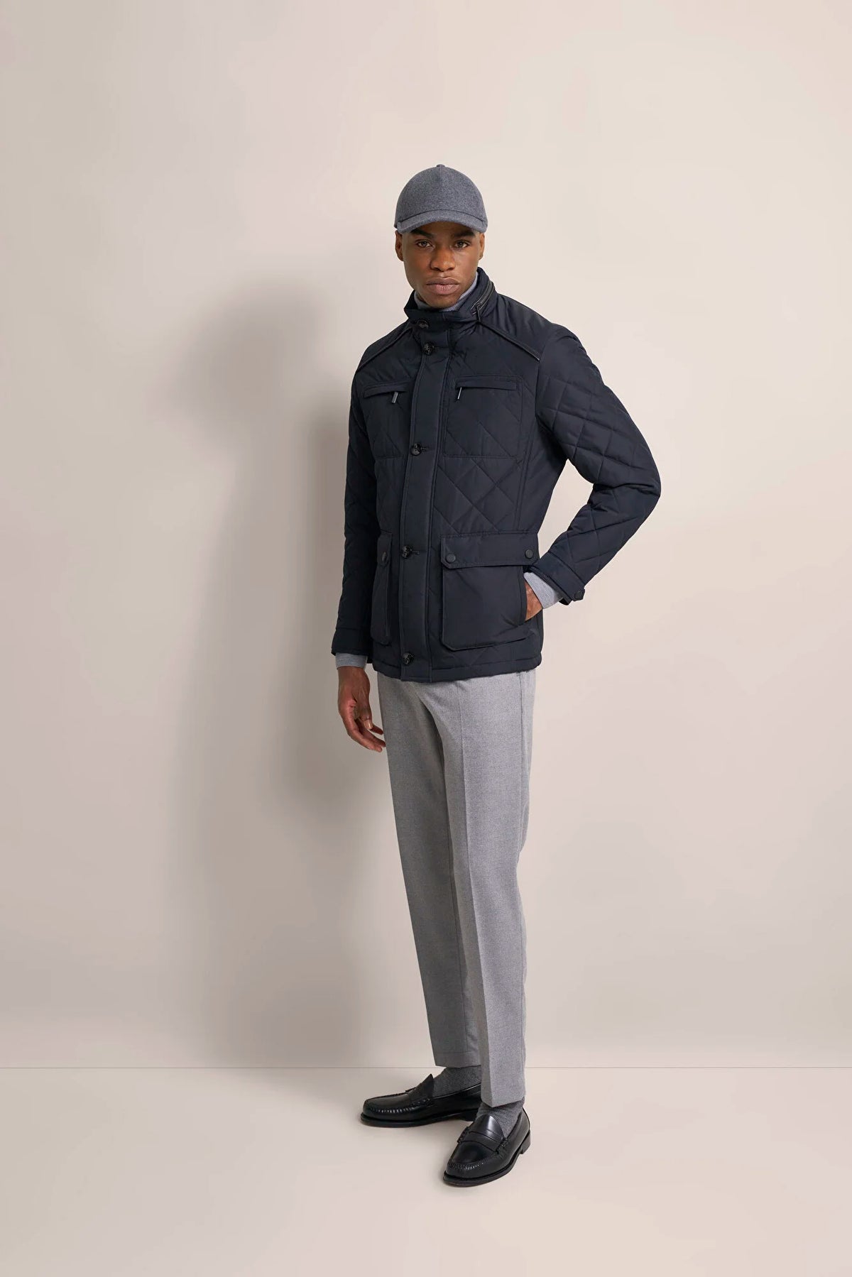 A man modeling a dark navy quilted jacket, light gray trousers, black loafers, and a gray baseball cap against a plain beige studio background.