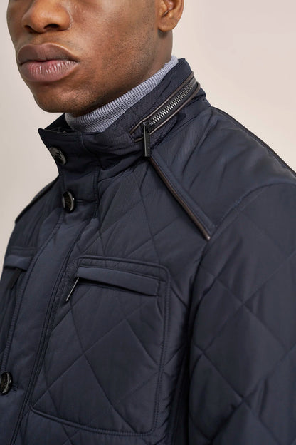 Close-up of a man wearing a navy quilted jacket over a light gray ribbed turtleneck, against a plain beige background.