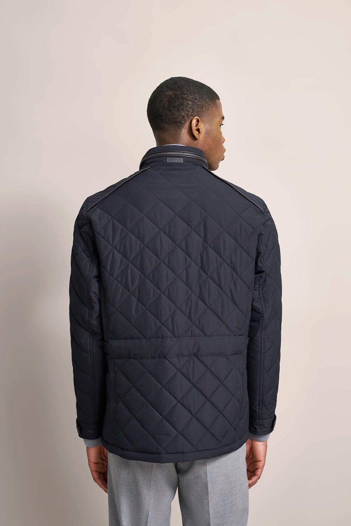 A man viewed from the back wearing a dark navy quilted jacket with a stand collar and zippered front, paired with light gray trousers, against a plain beige studio background.