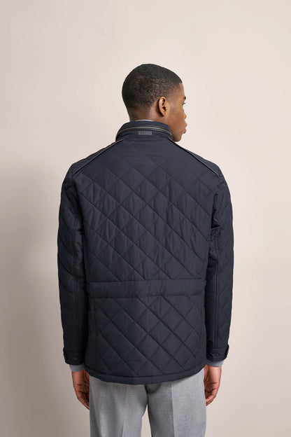 A man viewed from the back wearing a dark navy quilted jacket with a stand collar and zippered front, paired with light gray trousers, against a plain beige studio background.