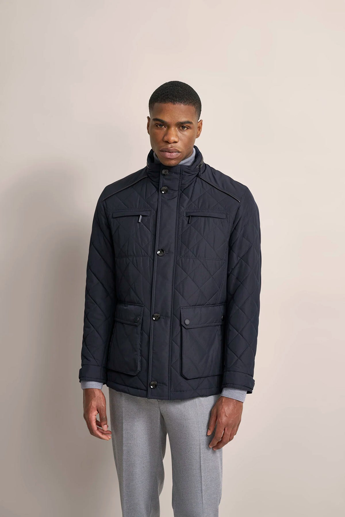 A man wearing a navy blue quilted jacket with a stand-up collar, front button closure, and four front flap pockets, paired with light gray trousers, standing against a solid beige studio background.