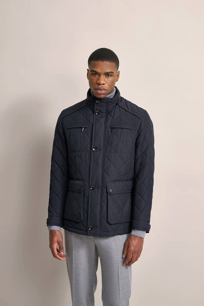 A man wearing a navy blue quilted jacket with a stand-up collar, front button closure, and four front flap pockets, paired with light gray trousers, standing against a solid beige studio background.