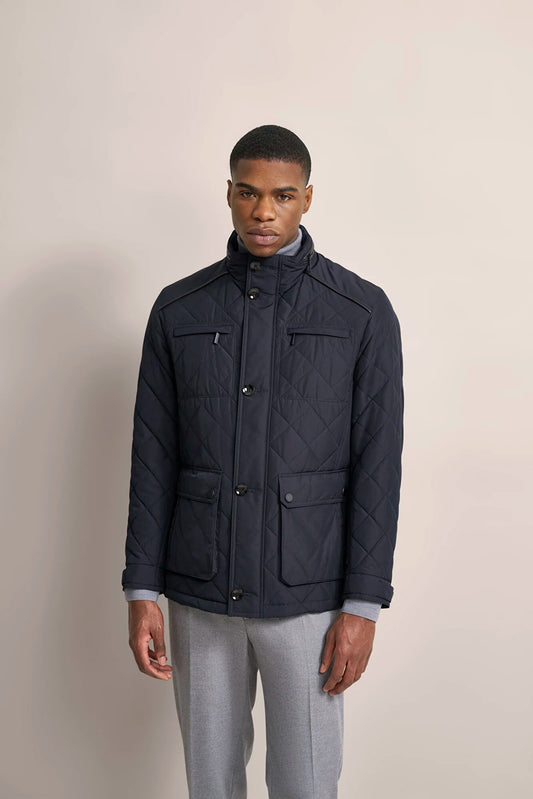 A man wearing a navy blue quilted jacket with a stand-up collar, front button closure, and four front flap pockets, paired with light gray trousers, standing against a solid beige studio background.